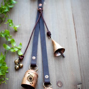 May include: Rustic hanging bell decoration featuring a dark brown leather strap with brass accents. The bells are a mix of sizes and shapes, including small round bells and larger, cylindrical bells with a sunburst design. The background is a wooden surface.