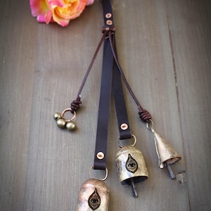 May include: A decorative leather strap with three metal bells and a cluster of small beads. The bells are gold-toned with an eye design. A pink and yellow rose rests nearby on a wooden surface.
