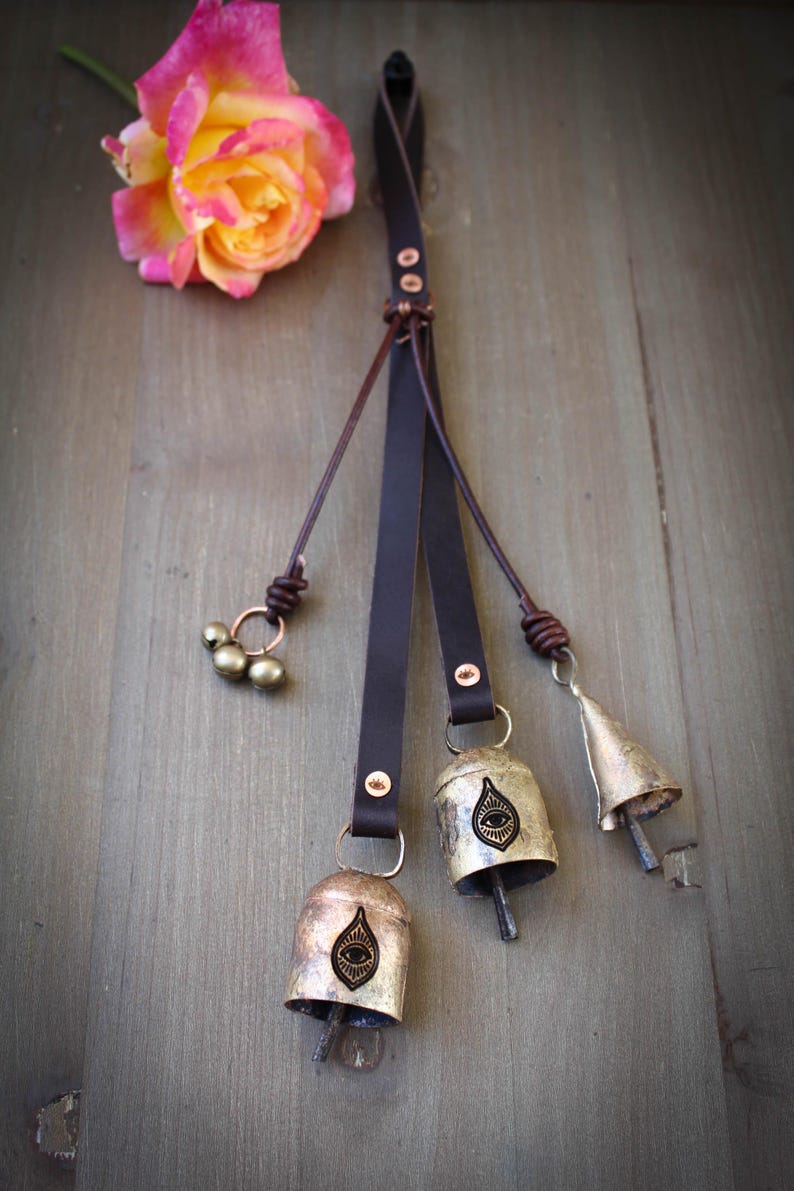 May include: A decorative leather strap with three metal bells and a cluster of small beads. The bells are gold-toned with an eye design. A pink and yellow rose rests nearby on a wooden surface.
