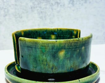 May include: A round, deep green ceramic yarn bowl featuring a glossy, mottled glaze with hints of yellow and blue. The bowl has a smooth, curved side with a vertical cutout slot for yarn to pass through, and it sits securely on a matching circular base.