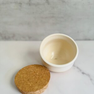 Puede incluir: Un pequeño recipiente de cerámica blanquecina con una tapa de corcho a juego. El recipiente es redondo con un borde ligeramente ensanchado. La tapa de corcho tiene forma circular plana. Los artículos están sobre un fondo blanco y gris.