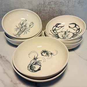 May include: A set of three off-white ceramic bowls stacked in groups. Each bowl features a detailed black line drawing of a different sea creature: one with a lobster, one with a crab, and one with two shrimp centered at the bottom of the basin.