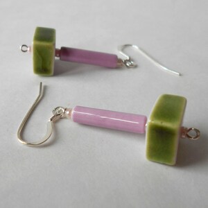 May include: A pair of handmade earrings featuring a green square bead, a pink cylindrical bead, and a small pink bead. The earrings have silver-colored ear wires. The earrings are displayed on a white surface.