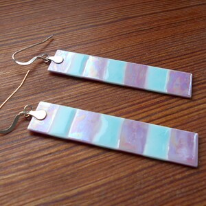 May include: A pair of iridescent earrings with a light blue and purple striped design. The earrings are made of glass and have silver hooks.