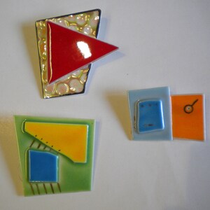 May include: Three ceramic tile brooches with geometric designs. The first brooch is a red triangle on a silver background with white dots. The second brooch is a green tile with a yellow triangle, a blue square, and gold lines. The third brooch is a blue tile with a smaller blue square inside, and a separate orange tile with a small circle.