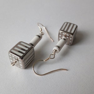 May include: A pair of silver earrings with white geometric ceramic cube pendants. The cubes have a striped pattern.