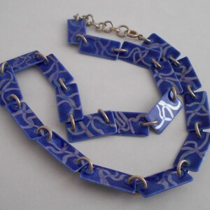 May include: A blue ceramic necklace with a silver chain and a white pattern. The necklace is made of rectangular links with a white design on a blue background.