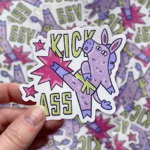 May include: A purple donkey with a yellow belt kicks its leg out in a karate pose. The donkey is surrounded by a pink starburst with glitter. The text "Kick Ass" is written in green letters.