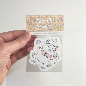 May include: A clear plastic bag with a white sticker on the front. The sticker has a cartoon illustration of a white cat with a pink bow and a crown. The text on the sticker reads "Bee and Puppycat".