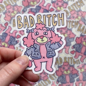 May include: A pink poodle wearing a blue denim jacket with a white cross on the back. The poodle has pink fur and a black nose. The sticker says "Bad Bitch" in yellow letters with a gold outline.