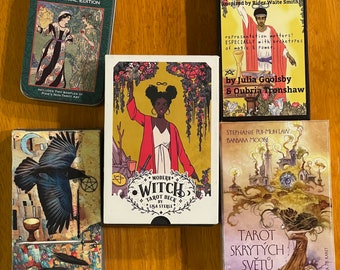 May include: Five different tarot decks, including the Smith-Waite Centennial Edition, Melaniated Classic Tarot, Modern Witch Tarot Deck, and Tarot Skrytych Svetu. The decks feature a variety of artistic styles and imagery.