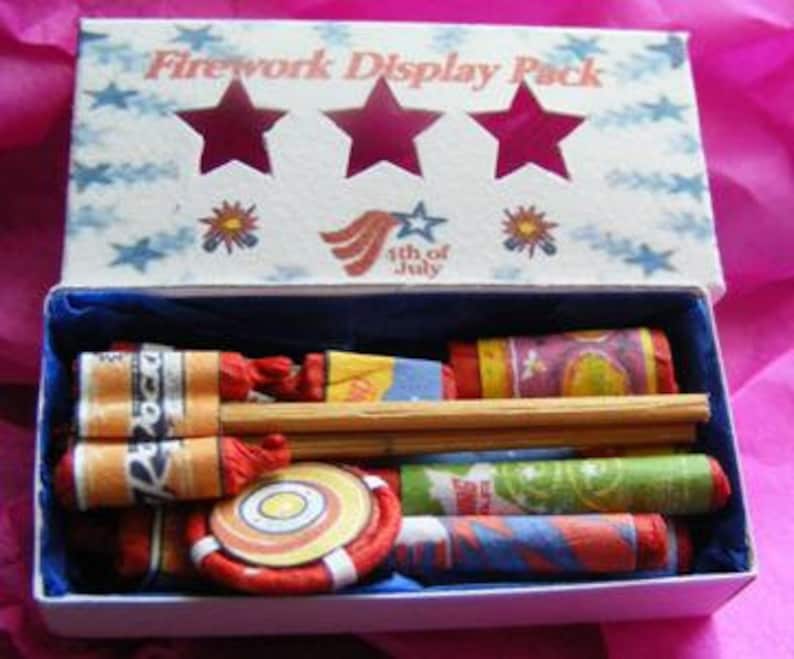 May include: A small box with a white background and blue stars. The box is open and contains a variety of small fireworks. The box is labeled "Firework Display Pack" and "4th of July".