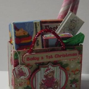 May include: A red and white gift bag with a handle, filled with wrapped gifts and candy canes. The bag has a label that says "Baby's 1st Christmas".