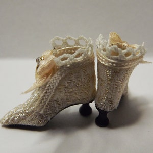 May include: A pair of white satin doll shoes with gold trim and lace detailing. The shoes have a small heel and are decorated with a small bow.