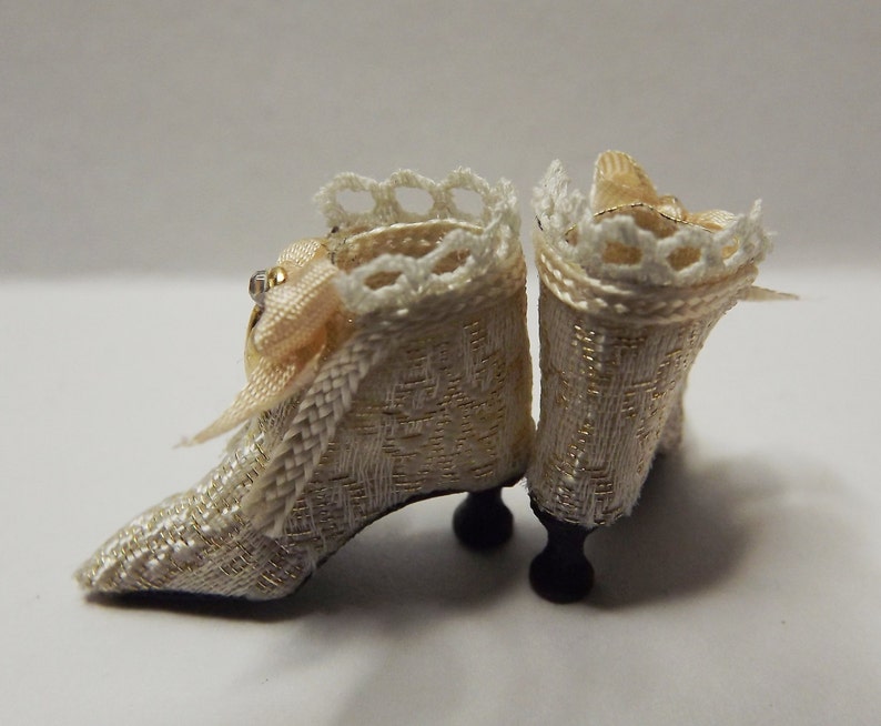 May include: A pair of white satin doll shoes with gold trim and lace detailing. The shoes have a small heel and are decorated with a small bow.