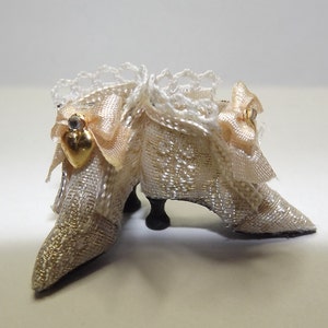May include: A pair of white satin doll shoes with gold trim and a heart-shaped embellishment. The shoes have a lace overlay and are decorated with a bow.