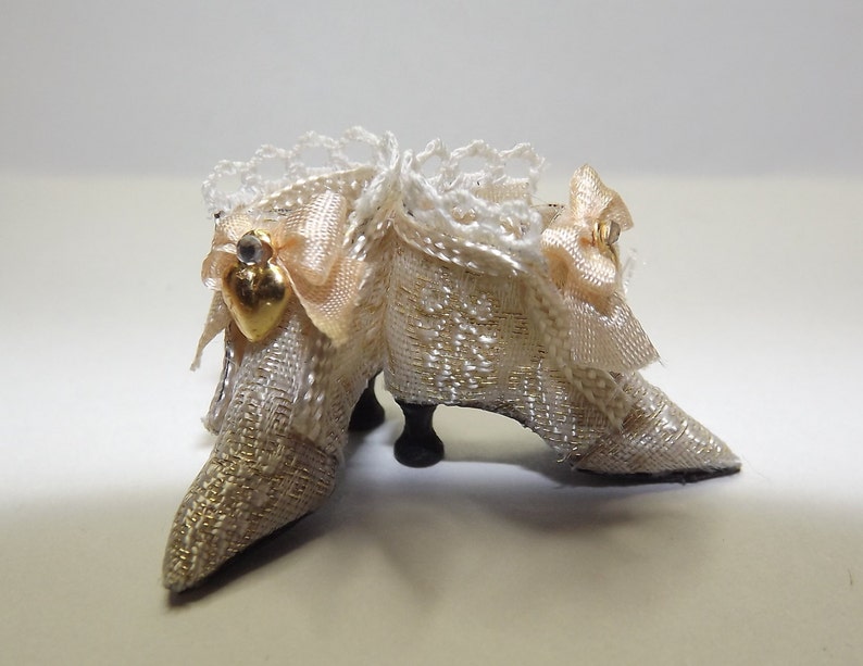 May include: A pair of white satin doll shoes with gold trim and a heart-shaped embellishment. The shoes have a lace overlay and are decorated with a bow.