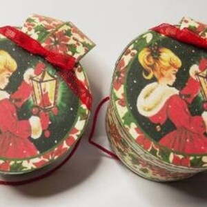 May include: Two round Christmas gift boxes with a red ribbon and a girl holding a lantern on each. The boxes are decorated with a green and red holly pattern.