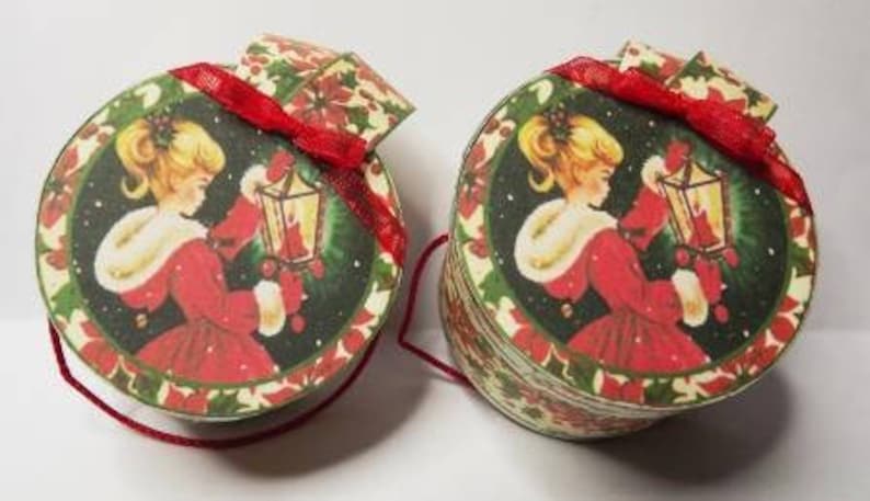May include: Two round Christmas gift boxes with a red ribbon and a girl holding a lantern on each. The boxes are decorated with a green and red holly pattern.