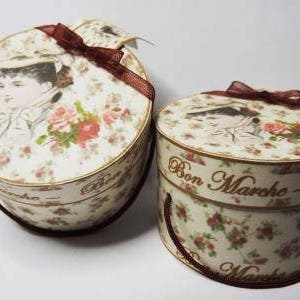 May include: Two cream-coloured, vintage-style round boxes with lids. They feature a floral pattern and a portrait of a woman on the lid. The smaller box has the text "Bon Marche". Each box is adorned with a brown ribbon.