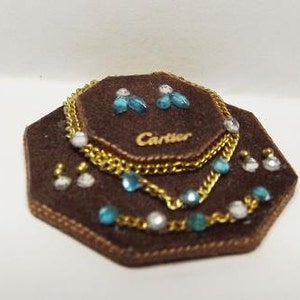 Op de afbeelding: Een bruin fluwelen juwelendoosje met een gouden kettingketting en oorbellen met blauwe en witte stenen. Het doosje heeft het woord "Cartier" erop gedrukt.