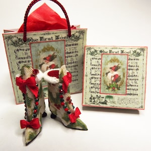 May include: Two miniature Christmas boots with red bows and holly accents. The boots are sitting in front of a small gift bag and box with a vintage Christmas design and the text "The First Noel".
