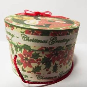 May include: A round, cream-colored gift box with a red ribbon and a red string handle. The box is decorated with a Christmas scene and the words "Christmas Greetings".