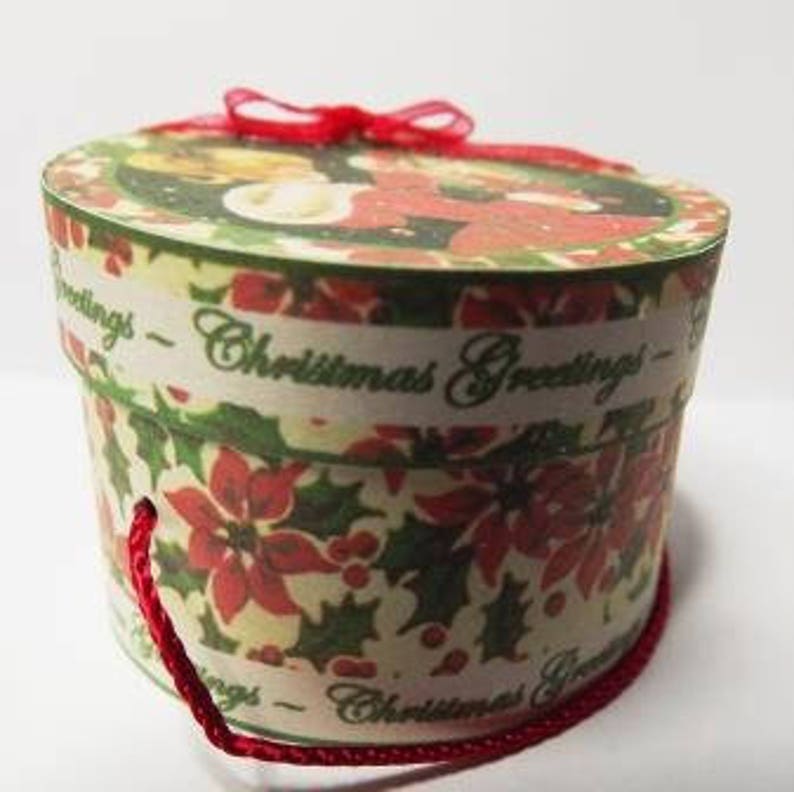 May include: A round, cream-colored gift box with a red ribbon and a red string handle. The box is decorated with a Christmas scene and the words "Christmas Greetings".