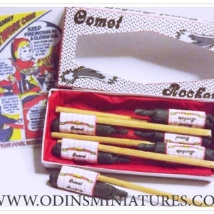 Op de afbeelding: Een rood-witte doos met een zwart-witte illustratie van een komeet. De doos bevat een set miniatuur vuurwerk met de labels "Comet" en "Rockats".