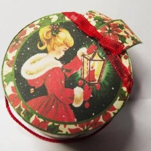 May include: A round, red and green Christmas ornament with a vintage illustration of a girl in a red dress holding a lantern. The ornament has a red ribbon attached to the top.