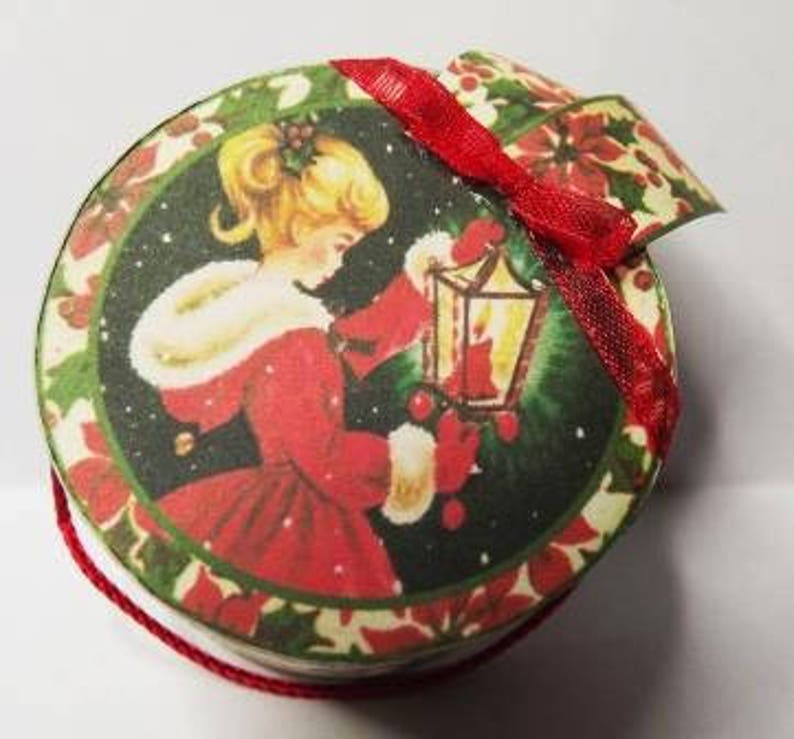 May include: A round, red and green Christmas ornament with a vintage illustration of a girl in a red dress holding a lantern. The ornament has a red ribbon attached to the top.