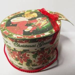 May include: A round, white paper gift box with a red ribbon. The box has a Christmas-themed design with a girl holding a birdhouse and the words "Christmas Greetings".