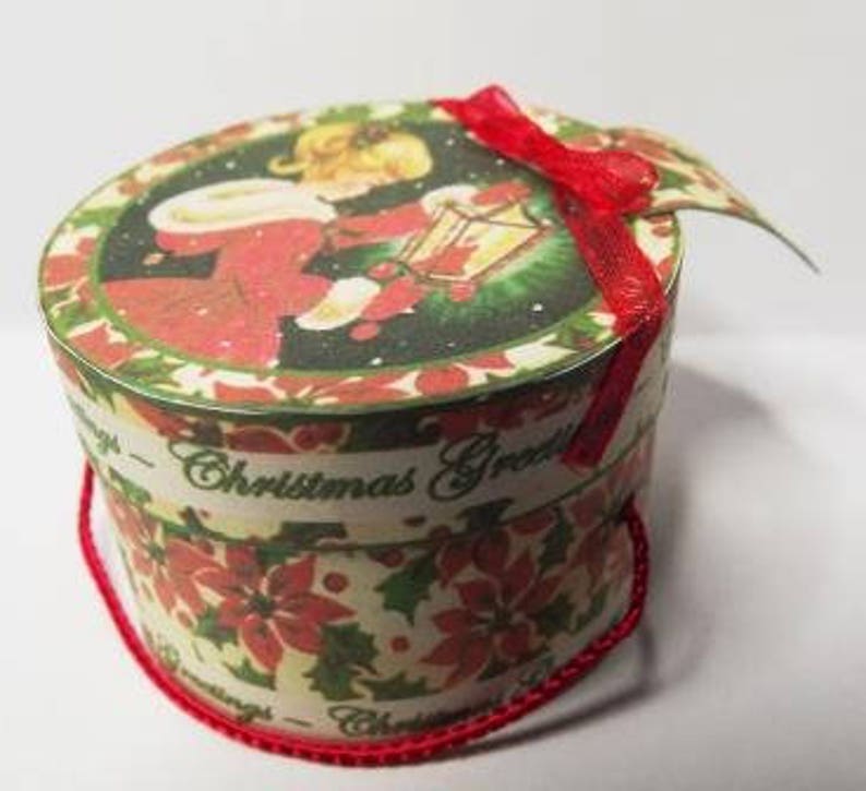 May include: A round, white paper gift box with a red ribbon. The box has a Christmas-themed design with a girl holding a birdhouse and the words "Christmas Greetings".