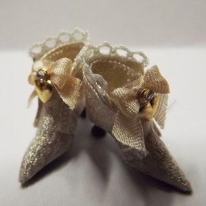 May include: A pair of white lace doll shoes with gold accents and a bow on each shoe.