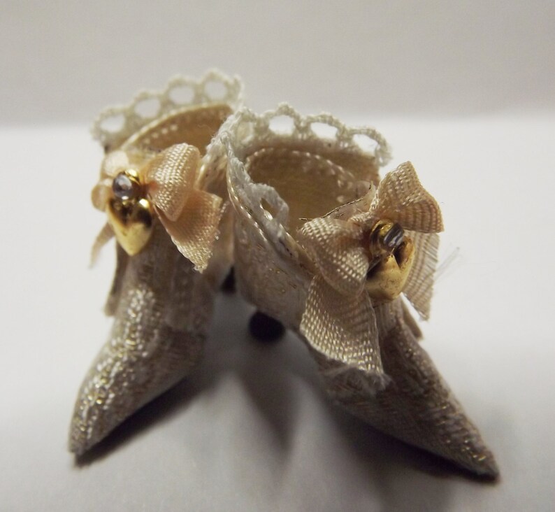 May include: A pair of white lace doll shoes with gold accents and a bow on each shoe.