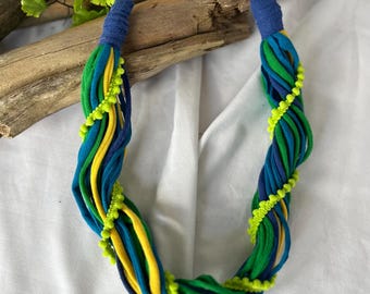 May include: A handmade necklace featuring a braided design with blue, green, and yellow fabric strands. The necklace is accented with neon green pom-poms and a blue fabric clasp. The necklace is displayed on a white surface.