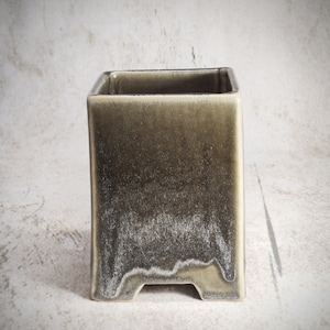 Può includere: Un vaso quadrato in ceramica con una smaltatura verde scuro e una finitura macchiettata. Il vaso ha quattro piccoli piedini sul fondo.