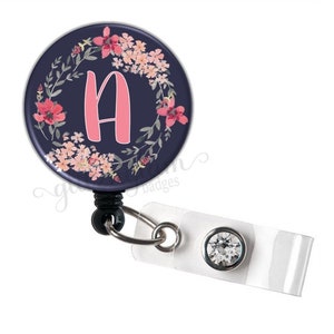 May include: A navy blue retractable badge holder with a floral wreath design and the letter "A" in pink. The badge holder has a clear plastic clip and a crystal-like button.