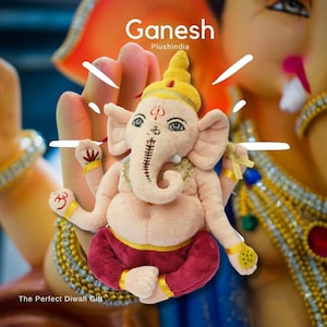 May include: A plush toy of the Hindu deity Ganesha, with a pink body, red pants, and a yellow crown. The toy is sitting with its legs crossed and its hands in a gesture of blessing. The background is a blurry image of a larger Ganesha statue.