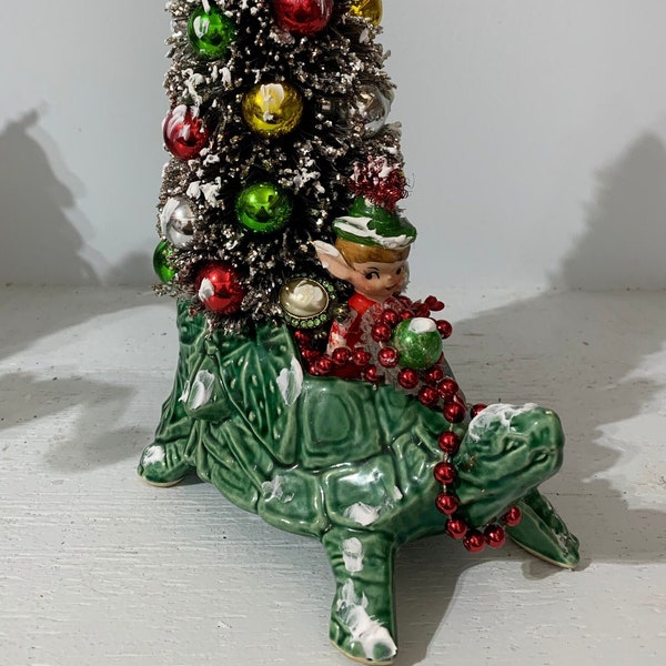 A vintage McCoy box turtle planter carrying a bottle brush Christmas tree being guided by a vintage elf.