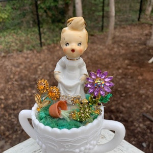 May include: A white ceramic cup with two handles, decorated with a raised grapevine design, filled with a miniature garden scene. The scene includes a small ceramic angel figurine, a tiny terracotta pot with a green plant, and various jeweled flowers and a frog.