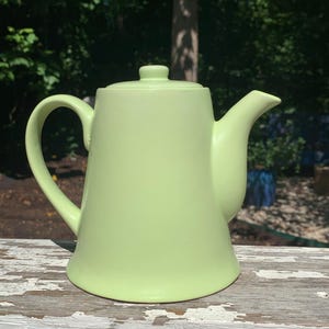 May include: A light green ceramic teapot with a curved handle, spout, and lid. The teapot is sitting on a weathered, white-painted surface, with a blurred green background of trees and foliage.