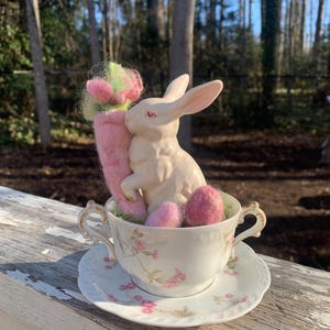 May include: A white ceramic bunny figurine holding a pink and green felt carrot, sitting in a vintage teacup with a floral pattern and pink felt eggs on a white saucer.