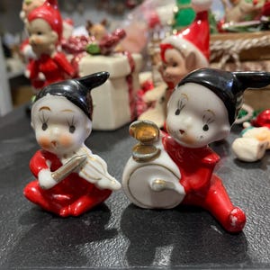 May include: Two vintage ceramic Christmas elf figurines. One elf is playing a violin and the other is playing a drum. Both elves are wearing red and white outfits with black hats.