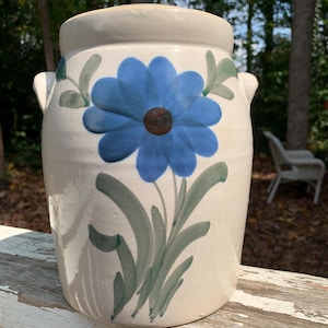 May include: Cream-colored ceramic jar with a large blue flower design. The flower has a brown center and is surrounded by green leaves and stems. The jar has two small handles and is sitting on a white surface.