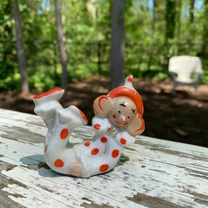 May include: A white porcelain clown figurine with red and orange polka dots, wearing a red hat with a white pom-pom. The clown is lying on its side with its arms outstretched.
