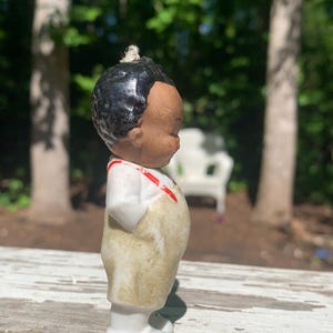 May include: A vintage porcelain figurine of a young child with black hair and a tan complexion. The figurine wears a white shirt with red suspenders and tan overalls. The figurine is standing on a white base.