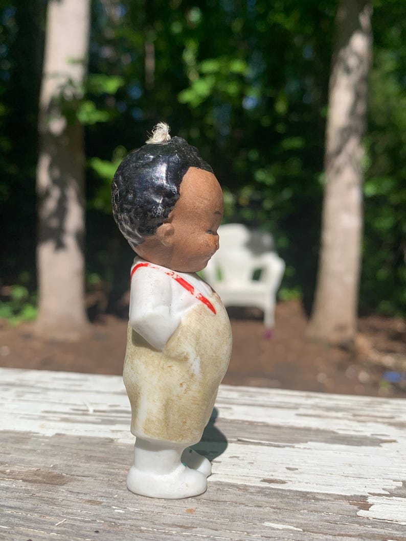 May include: A vintage porcelain figurine of a young child with black hair and a tan complexion. The figurine wears a white shirt with red suspenders and tan overalls. The figurine is standing on a white base.