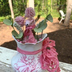 May include: A pink porcelain vase with a floral pattern and a pink ribbon. The vase is filled with pink flowers and a pink bunny figurine.