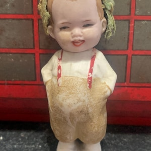 May include: A porcelain doll with a painted face and green hair. The doll is wearing a brown and white outfit with red straps.